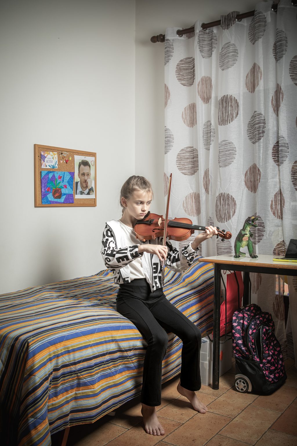 Sacrofano (Roma), 22 ottobre 2022. Slaviana Plakhotina (11 anni) studia per l’esame di violino che ha sostenuto on line con la sua scuola di musica di Odessa. Slaviana alloggia insieme alla sua famiglia in una casa messa loro a disposizione dalla parrocchia di Sacrofano, in provincia di Roma. La famiglia Plakhotina è composta da Yevgen (38), Oleksandra (34) e i figli Denys (14 anni, adottato all’età di 10 anni), Slaviana (11) e Savilii (3), nella .
La famiglia, originaria di Mariupol, viveva ad Odessa dal 2019. Yevgen (che in quanto padre di 3 figli è esonerato dalla legge marziale) lavora come marinaio per una compagnia marittima ucraina, mentre Oleksandra, dopo i suoi studi da giornalista, ha lavorato come corrispondente per una emittente di Mariupol e per un magazine ad Odessa. Ora si occupa a tempo pieno della casa e dei figli e scrive poesie. 
Il 24 febbraio, all’inizio dell’invasione russa, Yevgen si trovava a bordo di un cargo al largo di Rotterdam, sulle rotta Olanda, Finlandia e Russia. Il suo telefono non prendeva il segnale, e lui è stato informato dell’inizio della guerra dal secondo ingegnere di bordo, che avendo una sim olandese, riusciva a connettersi. Yevgen ricorda di aver pensato inizialmente ad uno scherzo, ma poi quando si è connesso tramite l’hotspot del collega, il suo telefono è stato subissato di messaggi. Il primo pensiero è stato ovviamente alla famiglia che però non è riuscito a contattare. Una volta in porto il suo telefono era regolarmente in funzione ed è riuscito a mettersi in contatto con Oleksandra, che nel frattempo si trovava insieme ai tre figli al sicuro in un bunker ad Odessa. Insieme hanno deciso che fosse più sicuro, per Oleksandra e i figli, lasciare Odessa. Oleksandra si aggrega ad altre due famiglie e con tre macchine si dirigono verso il confine con la Moldavia. Per arrivare al confine, distante neanche 100 km, hanno impiegato 18 ore, dormendo in auto. Nella sua Oleksandra, aveva spento anche il risc