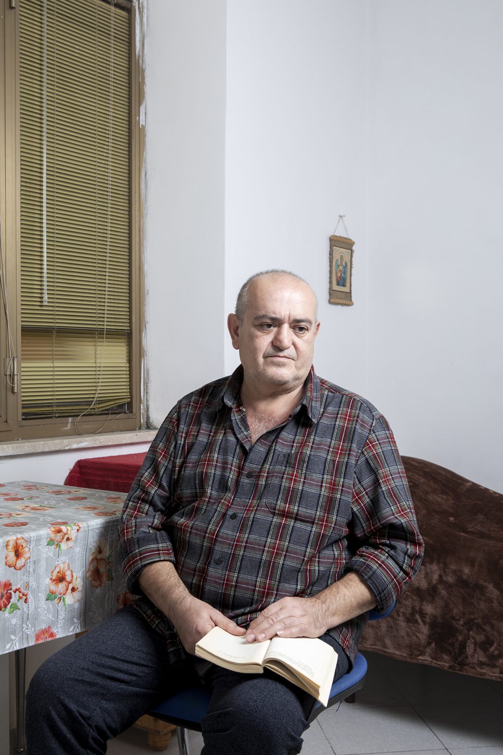 Anagni (FR), Italia, 15 Dicembre 2022. Serhii Avanesian, 54 anni, ritratto nel Centro di Accoglienza Straordinaria (CAS) gestito dalla cooperativa Diaconia in Anagni (FR) dove è attualmente ospitato insieme ad altri rifugiati ucraini. Serhii è nato in Azerbaijan da genitori di origine armena ed è un medico specialista della colonna vertebrale. Nel 1991 si è laureato in medicina all’università di Baku e nel 1992 ha dovuto lasciare la sua casa a Nagorno-Karabach a causa del conflitto tra la maggioranza etnica armena, sostenuta dalla Repubblica Armena, e la repubblica dell’Azerbaijan. Insieme alla moglie, anch’essa medico, si è trasferito a Pjatigorsk, in Russia. Dopo circa 10 anni, in seguito alla separazione dalla moglie, decide di trasferirsi a Kherson, in Ucraina. Li inizia a lavorare nell’ospedale cittadino e nel suo studio privato. Serhii riferisce di aver guadagnato bene, tanto che si è potuto permettere di costruirsi una villetta a circa 30 km da Kherson, dove viveva fino all’inizio del conflitto. La sua casa si trova sulla riva sinistra del fiume Dnipr, in un territorio attualmente occupato dall’esercito russo, proprio sulla strada percorsa dall’esercito russo proveniente dalla Crimea. Per due mesi ha vissuto sotto l’occupazione russa. Grazie alle sue disponibilità economiche all’inizio del conflitto era riuscito a farsi una cospicua scorta di generi alimentari e di prima necessità, che gli hanno permesso, durante il primo mese di occupazione, di non uscire di casa. Ha iniziato anche a farsi il pane da solo, che condivideva con gli anziani del luogo e altre persone bisognose. Racconta che per tutto il periodo dell’occupazione i vetri della casa tremavano incessantemente a causa dei rumori delle esplosioni e degli elicotteri che volavano a quota bassissima. Dopo il primo mese ha cominciato ad uscire di casa per procurarsi i viveri. Racconta che chi volesse uscire doveva esporre una bandiera bianca dall’automobile, precauzione che